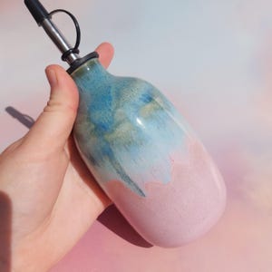 Op de afbeelding: Een keramische olieflacon met een roestvrijstalen tuit. De fles is beschilderd met een kleurengradiënt, van blauw en groen aan de bovenkant naar roze aan de onderkant. De fles wordt door een hand vastgehouden.