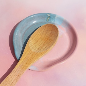 Könnte beinhalten: Ein Holzlöffel liegt auf einer Keramik-Löffelablage. Die Löffelablage ist eine Mischung aus Rosa und Blau, mit einem geschwungenen Design zum Halten von Utensilien. Der Holzlöffel ist hellbraun.