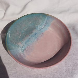 Può includere: Una ciotola in ceramica fatta a mano con un design bicolore. L'interno della ciotola è diviso diagonalmente, con una sezione turchese e blu da un lato e un rosa tenue dall'altro. Il bordo è rosa scuro.
