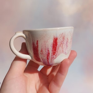 Puede incluir: Una taza de cerámica blanca con un asa negra y el texto "But First, Coffee" en un estilo manuscrito. La taza está sobre una superficie blanca. La taza mide 10 cm de alto.