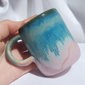 Könnte beinhalten: Eine handgefertigte Keramik-Tasse mit einem einzigartigen Farbverlauf. Die Tasse wechselt von einem dunkelgrünen Rand zu Blau- und Türkistönen und endet mit einem rosa Sockel. Der Griff hat einen goldenen Akzent. Ideal für Kaffee oder Tee.