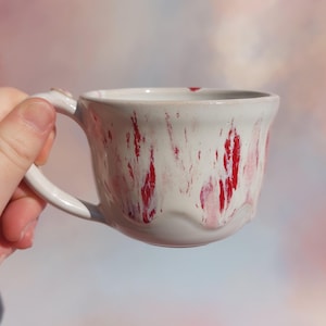 Op de afbeelding: Een witte keramische mok met een oor, versierd met rode strepen. De mok heeft een onregelmatige vorm, waardoor hij een handgemaakt gevoel krijgt. De mok wordt door een hand vastgehouden.