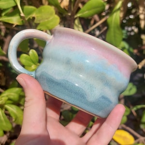 Pode incluir: Uma caneca de cerâmica com corpo arredondado e ondulado e uma única alça. O esmalte apresenta um gradiente que vai de um rosa pastel suave na borda, misturando-se com um azul celeste claro no centro e escurecendo para um azul-petróleo na base.