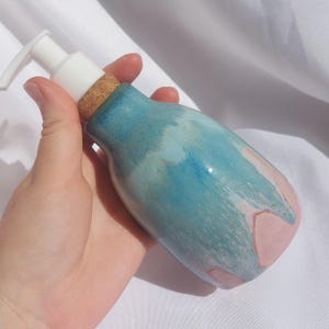 Op de afbeelding: Een keramische zeepdispenser met een witte pomp en een kurken basis. De fles is beschilderd met een mix van blauwe en roze tinten. De dispenser is ontworpen voor vloeibare zeep of lotion.