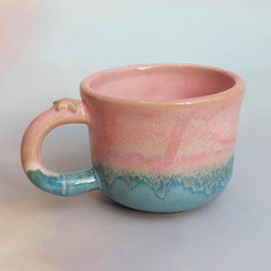 Può includere: Una tazza in ceramica fatta a mano con smalto rosa e blu. La tazza ha una forma arrotondata con un manico e un piccolo elemento decorativo. Il fondo della tazza è blu e la parte superiore è rosa.