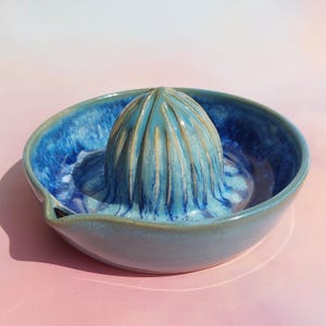May include: A handmade ceramic citrus juicer in shades of blue. The juicer features a ridged reamer in the center, surrounded by a bowl with a pouring spout. The glaze has a marbled effect, adding depth to the design.