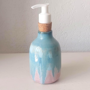 Könnte beinhalten: Ein Keramik-Seifenspender mit glänzender Oberfläche in einem Farbverlauf von Hellblau und zartem Rosa. Die Flasche hat einen zylindrischen Körper mit abgerundeter Schulter und einen Korkkragen am Hals, gekrönt von einer weißen Kunststoffpumpe.