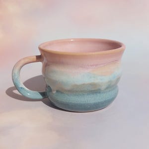 Peut inclure: Une tasse en céramique avec une forme arrondie et une anse lisse. L'extérieur présente un vernis dégradé passant d'un rose pastel doux au niveau du bord à un bleu clair atténué et un bleu sarcelle plus profond à la base, avec une finition brillante.