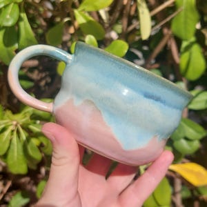 Pode incluir: Uma caneca de cerâmica com corpo arredondado e alça curva. O exterior tem um acabamento de esmalte bicolor, exibindo uma base rosa pastel suave que transita para um padrão de gotejamento azul claro perto da borda e na alça.