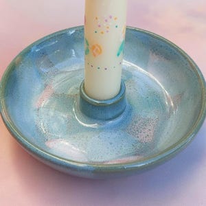 Può includere: Un portacandele in ceramica fatto a mano con una glassa azzurra. Il portacandele ha una forma a piatto poco profondo con un'area centrale rialzata per contenere una candela. È inserita una candela bianca con disegni floreali.