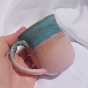 Peut inclure: Une tasse en céramique avec un design à deux tons. La partie supérieure de la tasse est d'un bleu sarcelle profond, tandis que la section inférieure et l'anse incurvée sont d'un rose pâle doux. La transition entre les deux couleurs est subtile.
