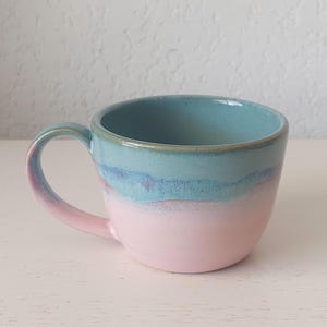 Peut inclure: Une tasse en céramique avec un émail bicolore, présentant une base rose pastel douce qui se transforme en une section supérieure bleu clair. La tasse possède un corps arrondi, une anse incurvée sur le côté gauche et une finition lisse et brillante.
