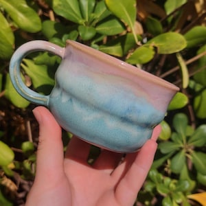 Peut inclure: Une tasse en céramique présentant une forme arrondie et organique avec une anse lisse. L'émail extérieur passe d'un rose pastel doux sur le bord supérieur à un bleu turquoise vif et moucheté à la base, créant un aspect mélangé et coloré.
