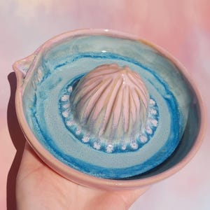 Op de afbeelding: Een handgemaakte keramische citruspers met een lichtroze ruimer en een turquoise kom. De kom heeft een lichtroze rand en een donkerblauwe ring. De citruspers wordt in een hand gehouden.
