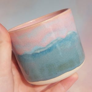 May include: A handmade ceramic mug with a gradient design. The top is a soft pink, transitioning to a vibrant blue-green at the base. The mug has a smooth, glossy finish and a natural, unglazed base.
