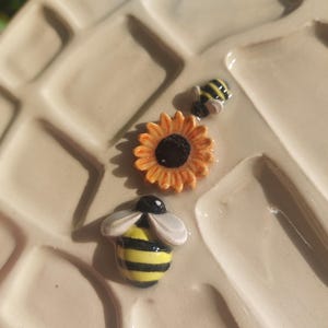 Può includere: Primo piano di un piatto in ceramica con superficie strutturata. Il piatto presenta tre piccoli pezzi decorativi in ceramica: un girasole con petali arancioni e centro nero, e due api con strisce gialle e nere.