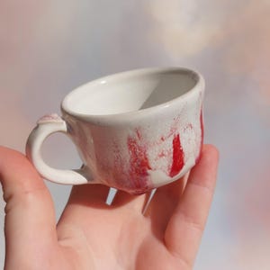 Puede incluir: Una pequeña taza de té de cerámica hecha a mano. El interior es blanco y el exterior presenta un diseño de vetas rojas sobre un fondo blanco. La taza tiene un asa pequeña y una forma irregular, lo que sugiere que está hecha a mano.