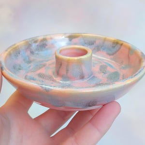 Può includere: Un portacandele in ceramica con base circolare e supporto cilindrico centrale. Il supporto è smaltato in una miscela di tonalità rosa, blu e oro. Il pezzo è tenuto in mano, mostrando le sue dimensioni e il suo design.