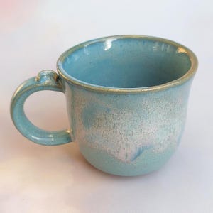 Può includere: Tazza in ceramica fatta a mano con smalto azzurro chiaro. La tazza ha una forma arrotondata, un manico e un bordo dorato. Lo smalto ha un aspetto macchiettato, che aggiunge consistenza. Perfetta per caffè o tè.