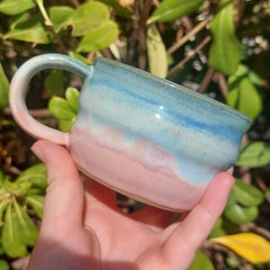 Pode incluir: Uma caneca de cerâmica com um corpo liso e arredondado com um gradiente de cor horizontal. A parte superior é vidrada num azul-celeste vibrante, que transita para um rosa pálido suave na base. Inclui uma pega curva com cores a condizer.