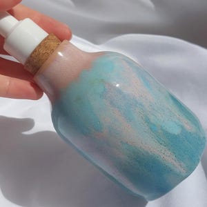 Könnte beinhalten: Ein Keramik-Seifenspender mit weißer Pumpe und Korken. Die Flasche hat eine blaue und rosa Glasur mit glänzendem Finish. Der Spender ist für Flüssigseife oder Lotion konzipiert.