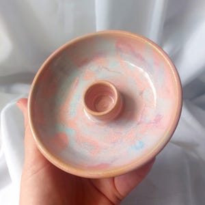 Può includere: Un portacandele in ceramica fatto a mano con un design circolare. Il portacandele presenta un cilindro centrale rialzato per la candela e un piatto largo e poco profondo. Il pezzo è smaltato in una tavolozza di colori pastello rosa, pesca e blu.