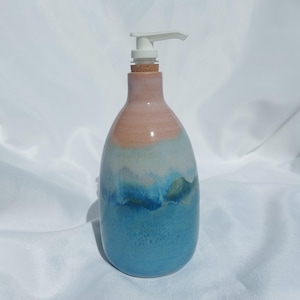 Op de afbeelding: Een keramische zeepdispenser met een afgerond lichaam voorzien van een kleurverloopglazuur dat overgaat van een zachte perzikkleur aan de bovenkant naar een diep oceaanblauw aan de basis. De dispenser heeft een witte plastic pomp en een kurken kraag.