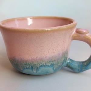 May include: A handmade ceramic mug with a pink interior and exterior, transitioning to a turquoise base. The handle and rim have a yellow-tan glaze. The mug is designed with a rounded shape.