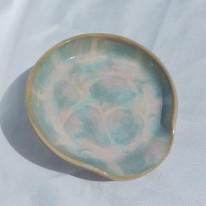Op de afbeelding: Een ronde keramische lepelhouder met een gemarmerd glazuurpatroon in zachte pasteltinten van lichtblauw en bleekroze. De randen van de schaal zijn afgewerkt met een natuurlijke, ongeglazuurde beige rand die contrasteert met de glanzende binnenkant.