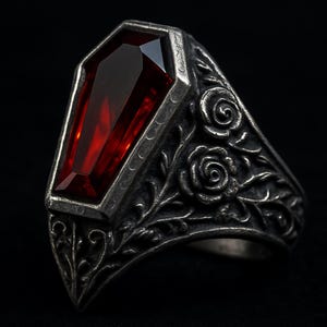 Puede incluir: Anillo de estilo gótico con una gema roja oscura en forma de ataúd. El anillo es de metal plateado y presenta intrincados detalles florales y de rosas en la banda. La gema está facetada, captando la luz y creando un efecto visual llamativo.