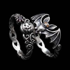 May include: Two silver rings featuring oxidized metalwork. One ring displays a carved jack-o'-lantern pumpkin with a dark red gemstone, while the other ring features a detailed bat with orange gemstone eyes and wings spread across the band.
