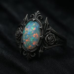 Puede incluir: Un anillo plateado con una piedra de ópalo ovalada. El anillo presenta detalles ornamentados, incluyendo rosas y puntas. El ópalo muestra un juego de colores con tonos azules, naranjas y verdes, sobre un fondo oscuro.