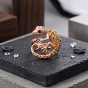 May include: A rose gold lizard ring with a ruby eye and a body encrusted with yellow and green gemstones. The ring is set on a black stone surface with scattered clear crystals.
