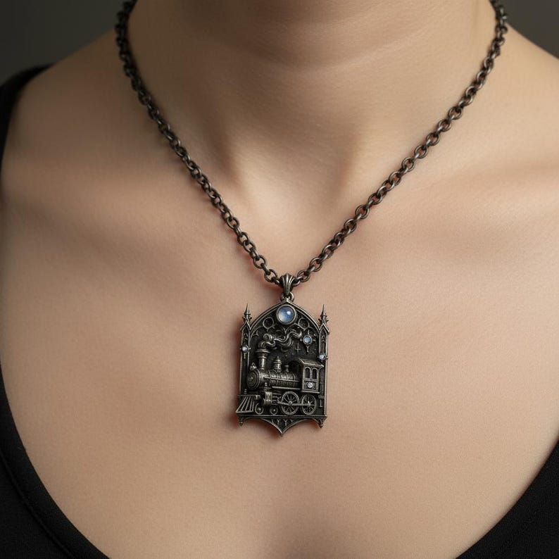 Puede incluir: Un collar con colgante plateado con un dise&ntilde;o detallado de tren. El colgante tiene forma rectangular con una parte superior arqueada, adornado con peque&ntilde;as gemas azules y una piedra lunar central. La cadena es de color plata oscura a juego.