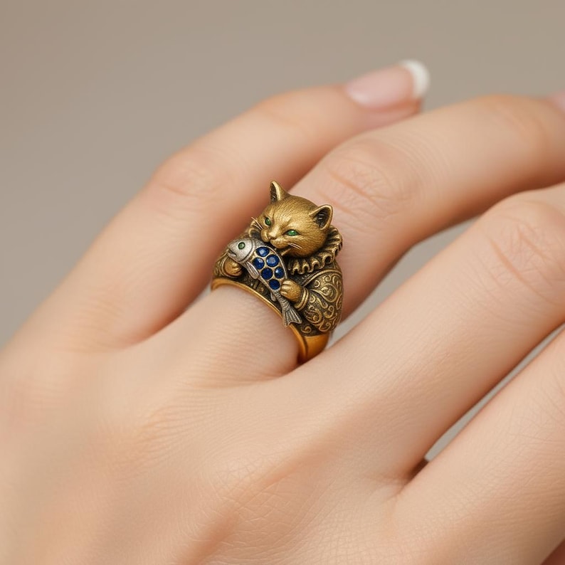 Puede incluir: Un anillo dorado con un gato sosteniendo un pez azul y plateado. El gato tiene ojos verdes y lleva un collar decorativo y ropa estampada. El anillo est&aacute; en un dedo.