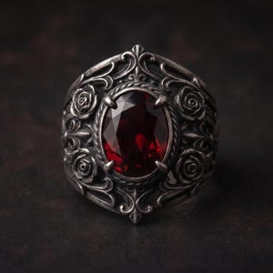 May include: A silver ring featuring an oval-cut deep red gemstone set in the center. The band is crafted with intricate oxidized metalwork, displaying detailed relief carvings of roses and scrolling filigree patterns surrounding the stone.