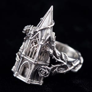 May include: A silver ring featuring a detailed architectural structure resembling a gothic cathedral tower. The band is adorned with sculpted rose vines, thorns, and blooming rose flowers that wrap around the central tower design.