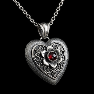 Peut inclure: Un pendentif en forme de cœur sur une chaîne en argent avec une texture martelée, un motif floral en filigrane complexe et une pierre précieuse rouge foncé facettée au centre. Le métal présente une finition oxydée soulignant les détails.