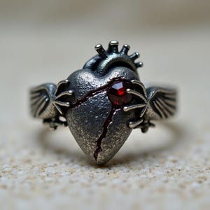 May include: A silver-toned ring featuring a detailed anatomical heart design with a crack and a red gemstone. The ring has small wings on each side of the heart.