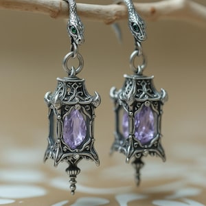 May include: A pair of silver-toned earrings featuring a lantern-shaped design with an intricate metal frame and a central faceted light purple gemstone. The top of each earring is shaped like a snake head with small green eyes and a hook.