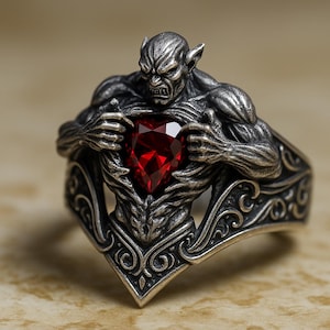 May include: A silver ring with a detailed, sculpted figure of a creature with a fierce expression. The creature holds a heart-shaped, faceted, deep red gemstone. The ring band has an ornate, swirling design.