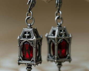Puede incluir: Un par de pendientes plateados con un gancho en forma de mano esquelética unido a un colgante con forma de linterna. Cada linterna tiene una piedra preciosa roja oscura facetada, un marco de metal y una pequeña cruz.