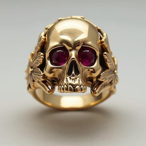 May include: A gold-colored skull ring with ruby-colored gemstones for eyes. The skull is surrounded by leaf-like details. The ring is a statement piece, perfect for those who appreciate unique jewelry.