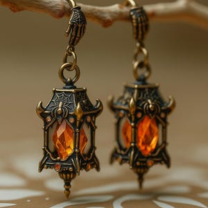 Op de afbeelding: Een paar oorbellen in antieke stijl met miniatuur lantaarnhangers. Elke lantaarn heeft een sierlijk bronskleurig frame met een spinnenwebontwerp, een grote oranje edelsteen en een handvormige haak. De oorbellen hangen aan een tak.