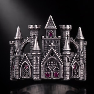 May include: A silver-toned ring shaped like a miniature castle with multiple towers, pointed spires, and arched windows. The structure is accented with small, round, deep red gemstones embedded within the masonry and window frames.