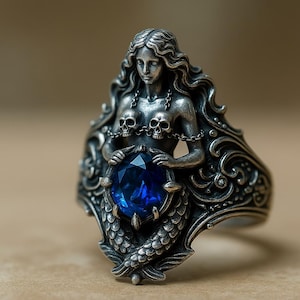 May include: A silver-toned mermaid ring featuring a large, oval, deep blue gemstone. The mermaid has long flowing hair, skull accents, and a detailed tail. The ring has an ornate, gothic design.