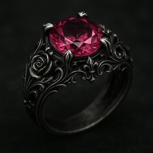 May include: A dark, ornate ring featuring a large, faceted pink gemstone. The ring has a detailed, floral design with roses and other decorative elements. The metal has a dark, antique finish, creating a gothic or vintage aesthetic.