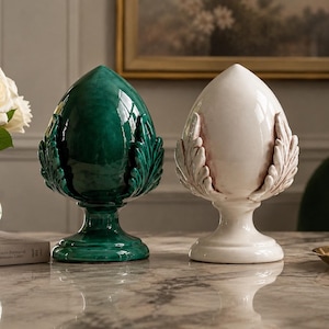 Peut inclure: Deux ornements décoratifs en céramique, un vert émeraude et un blanc, sont posés sur une surface en marbre. Chacun a un motif en forme de feuille autour de la base et un sommet lisse en forme d'œuf. Un vase de roses blanches et un bougeoir sont également présents.