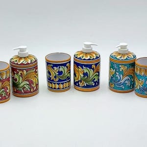 Puede incluir: Conjunto de cuatro accesorios de baño de cerámica con diseños florales. El conjunto incluye dos dispensadores de jabón y dos vasos. Los dispensadores tienen bombas blancas y los vasos están abiertos en la parte superior. Los diseños presentan patrones florales azules, verdes, amarillos y rojos sobre un fondo blanco.