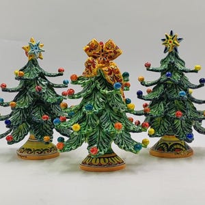 以下が含まれることがあります： 3つのセラミッククリスマスツリーの置物。木は緑色で、カラフルなオーナメントと星またはリボンのトッパーが付いています。ベースには装飾的な模様があります。木は高さ約15cmです。